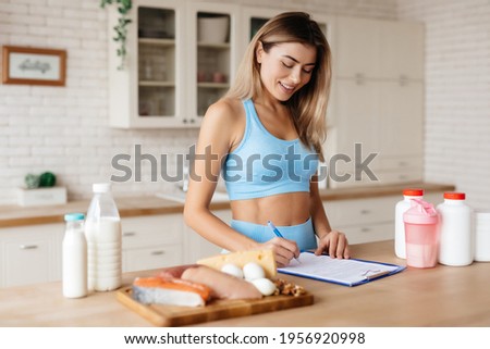 Attractive lady taking notes and standing near kitchen table with meat, dairy products and food supplements on it