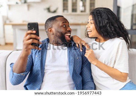Smiling millennial black couple sit on couch in kitchen have fun browsing Internet on modern smartphone together, happy young husband and wife relax on sofa use cellphone enjoy family weekend at home