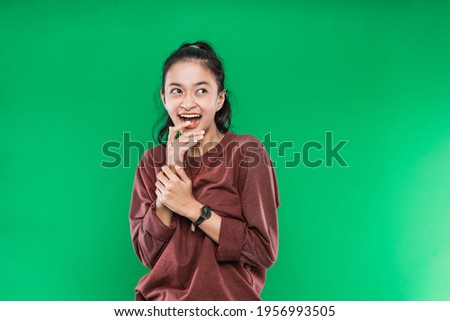 young Asian woman expressing happy wide-open mouth looking beside and raising hands on the chin on green background