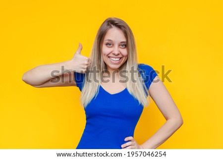 A young caucasian beautiful happy cheerful smiling blonde woman in a blue t-shirt showing thumb up like gesture with her hand isolated on a bright color yellow background. Positive human emotion