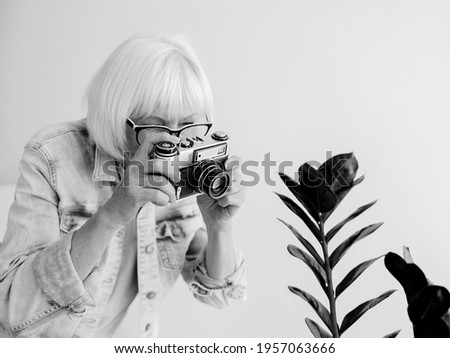 monochrome portrait of senior stylish woman with gray hair and in glasses and denim jacket taking pictures with film camera. Age, hobby, anti age, positive vibes, photography concept