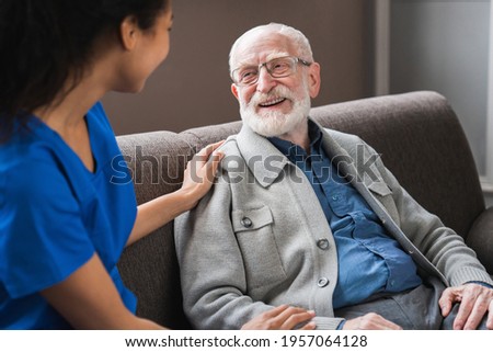 Young friendly african female caregiver talking chatting to happy senior man in hallway of nursing home. Picture of smiling nurse assisting senior elderly man Royalty-Free Stock Photo #1957064128