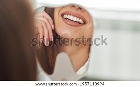 Happy young woman smiling checking out her perfect healthy teeth in the mirror close up, at the dentist office Royalty-Free Stock Photo #1957110994