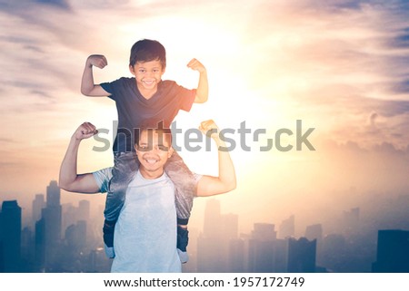 Young father piggybacking his son while showing their biceps with sunlight background Royalty-Free Stock Photo #1957172749