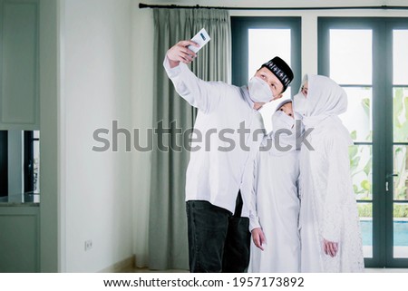 Happy Muslim family wearing face mask while using a smartphone to taking a selfie photo together during Eid Mubarak in the living room at home