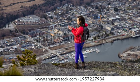 Adventurous Female Hiker on top of Chief Mountain taking pictures of a beautiful view from above of Canadian Nature and Touristic Town. Located in Squamish, North of Vancouver, BC, Canada.