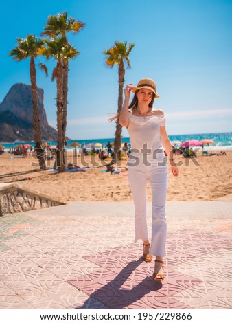 A Caucasian redhead dressed in white and with a straw hat walking along the beach of Calpe in summer, Valencia. Spain