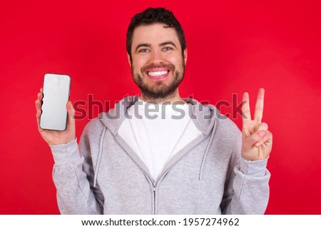 young handsome caucasian man in sports clothes against red background holding modern device showing v-sign