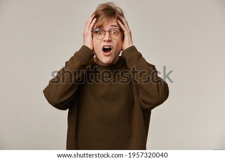 Teenage guy, terrified looking man with blond hair. Wearing brown sweater, glasses and has braces. Holds his head in fear. Emotion concept. Watching at the camera isolated over grey background