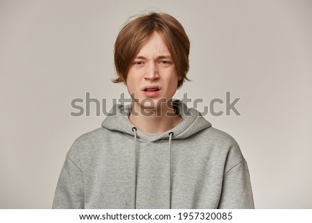 Irritated looking male, displeased guy with blond hair. Wearing grey hoodie. Has braces. People and emotion concept. Frowns his face and watching at the camera isolated over grey background
