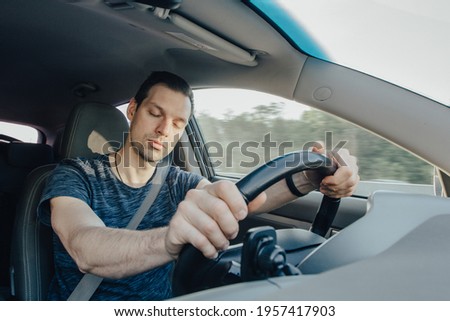 Fatigue of sleeping driver driving at speed while holding wheel while driving on highway in forest. Front view of exhausted man. Crisis or stress of person during road trip. Royalty-Free Stock Photo #1957417903