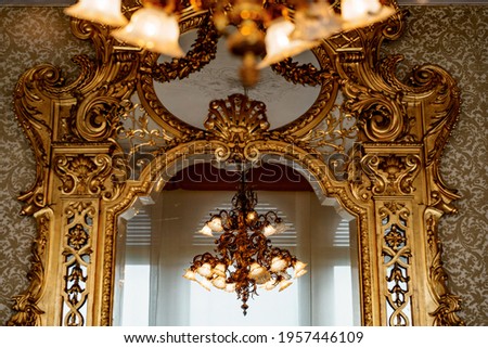 A large gold chandelier with patterns and many shades is reflected in a large antique wall mirror with a gilded carved frame.