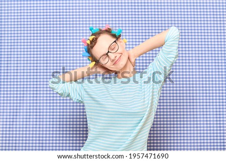 Portrait of happy blond girl with bright colorful hair curlers on head, smiling, with eye closed from pleasure, stretching hands behind head, standing over shower curtain background. Beauty concept