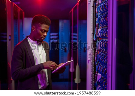 Serious young African-American data engineer standing at open server rack cabinet and using tablet while setting up system at data center Royalty-Free Stock Photo #1957488559