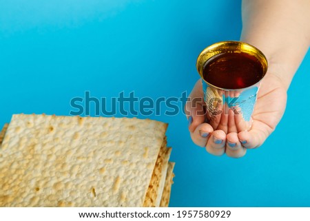 Plates of matzo laid on top of each other on a blue background and a glass of kiddush wine in hand.
