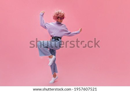 Happy young woman in stylish jeans and purple sweater dance on pink background. Curly short-haired girl move in great mood on isolated background . Royalty-Free Stock Photo #1957607521