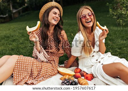Tanned happy brunette woman in boater and beige polka dot outfit sits on white rug outside. Happy excited blonde lady in sunglasses has picnic with friend and holds piece of melon. Royalty-Free Stock Photo #1957607617