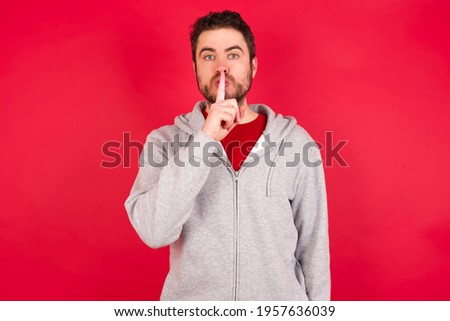Young caucasian man wearing tracksuit over red background makes silence gesture, keeps finger over lips. Silence and secret concept.