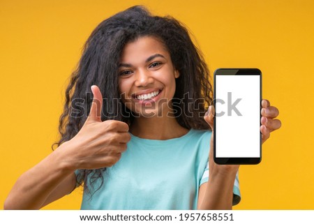 Cute african american girl with perfect smile holding mobile phone demonstrating blank white screen directly at the camera and holding thumb up. Mock up, copy space for your text. Focus is on hands. 