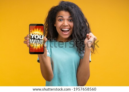 Beautiful smiling dark skinned woman showing sincere excitement about victory at bookmaker's website. Girl being happy winning bet in online sport gambling application on her mobile phone.