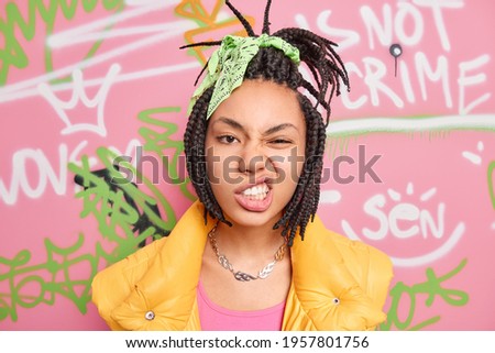 Teenage lifestyle concept. Cheeky hipster girl with combed dreadlocks clenches teeth smirks face looks self confident at camera spends free time at street poses over graffiti wall background
