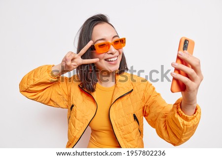 Beautiful stylish Asian woman takes selfie on mobile phone makes v sign smiles at camera has positive face wears orange sunglasses and jacket isolated over white background. Modern lifestyle