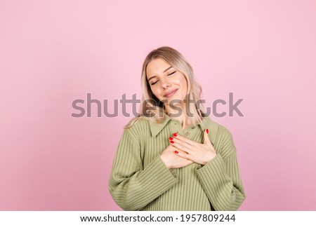 Pretty european woman in casual sweater on pink background holding hands folded on chest, heart copy space