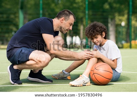 Side view portrait of focused adult man coach helping boy with knee trauma after playing basketball on the court, sad player feeling pain, touching leg. Activity And Sports Injury Concept. Royalty-Free Stock Photo #1957830496