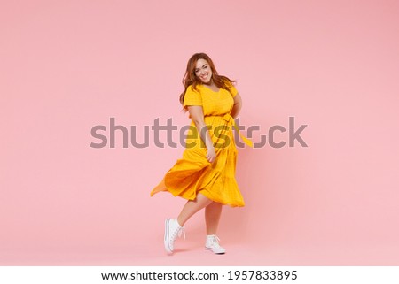 Full length portrait of smiling beautiful charming young redhead plus size body positive female woman girl 20s in yellow dress posing looking camera isolated on pastel pink color background studio Royalty-Free Stock Photo #1957833895