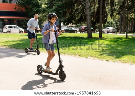 Happy Loving Family. Full body length photo of excited curly boy in protective helmet driving motorized push scooter with daddy at park, spending time together outdoors at summer, selective focus Royalty-Free Stock Photo #1957841653