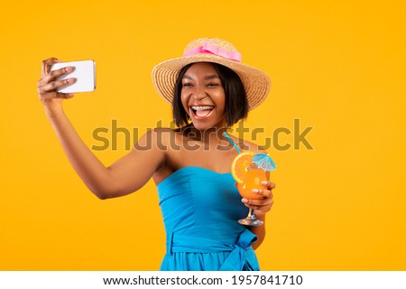 Happy black woman with yummy summer cocktail taking selfie on smartphone over orange studio background. Sweet African American lady with refreshing beverage making photo of herself