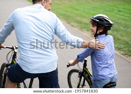 Healthy Family Relationship. Rear back view portrait of smiling father and son riding bicycles down the street in public park, man tapping and patting boy on the shoulder, teaching and showing support Royalty-Free Stock Photo #1957841830