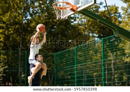 Happy Loving Family. Portrait of young curly boy sitting on father's shoulders, playing basketball, throwing ball to basket, man standing on court outdoors and helping his son, selective focus Royalty-Free Stock Photo #1957850398
