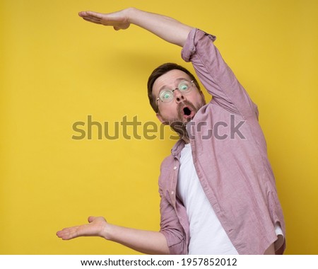 Surprised man in glasses shows a large size with hands, he exaggerates and looks at the camera in amazement. Copy space.  Royalty-Free Stock Photo #1957852012