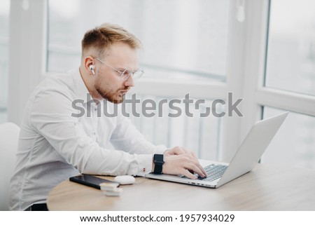young man in glasses wearing wireless headphones works with laptop, video conference Royalty-Free Stock Photo #1957934029