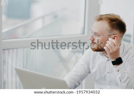 young man in glasses wearing wireless headphones works with laptop, video conference Royalty-Free Stock Photo #1957934032
