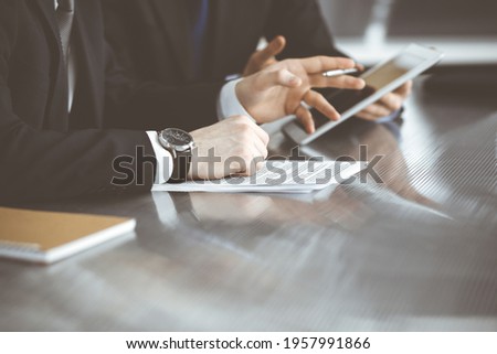 Unknown businessmen using tablet computer and work together at the glass desk in modern office, close-up. Teamwork and partnership concept