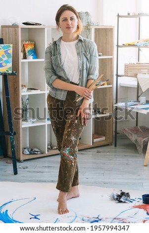 Art class. Painter lifestyle. Painting skill. Creative leisure. Confident relaxed female artist with paintbrushes standing on colorful stained abstract artwork on floor in light modern studio.