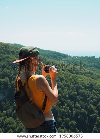 A girl in a cap takes pictures on the phone of the green surroundings of a mountain valley on a clear summer day