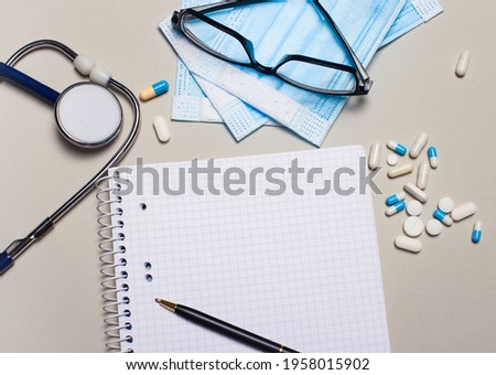 On a light gray background, a stethoscope, pills, face mask and glasses. Nearby is a pen and a blank notebook with space to insert text. Medical concept. Template