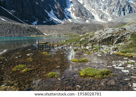 Beautiful scenic landscape with turquoise mountain lake with transparent water and stony bottom. Azure glacial lake with clear water surface in sunlight. White snow and green grass near mountain lake.