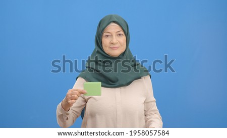 The old woman in a turban holding a green box in her hand points to the box with her finger and makes a thumbs up. Studio shot isolated on blue background. 