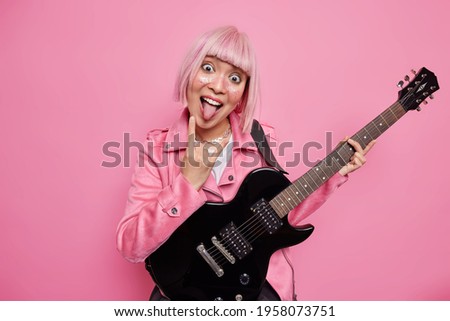 Energetic female celebrity makes rock n roll gesture sticks out tongue being passionate about music foolishes around while playing electric guitar has large audience poses indoor isolated on pink