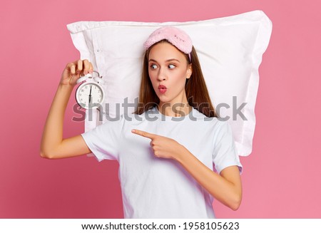 Funny young woman with pillow pouting lips and pointing at alarm clock after awakening against pink background in morning