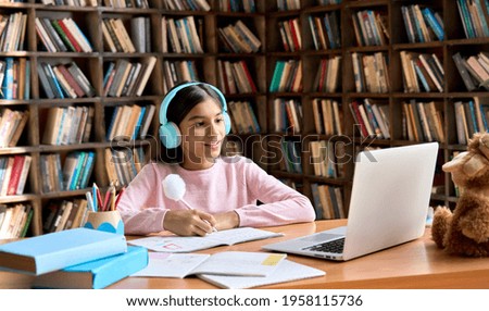 Happy smiling indian junior student wearing headphones having online virtual learning language class on laptop. Cute latin schoolgirl writing in workbook watching video meeting with teacher.