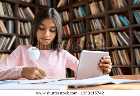Concentrated arab indian girl student doing homework with learning online video class webinar in virtual classroom on digital tablet device. Hispanic schoolgirl writing in notebook. Study from home.