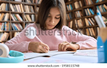 Adorable cute indian latin schoolgirl studying at home sitting at table. Serious spanish arab pretty kid junior student learning writing in work book doing homework at home classroom.