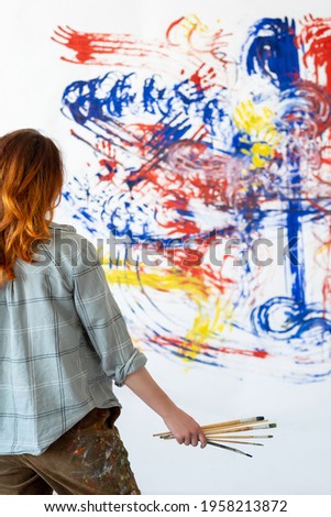 Finger painting art. Interior decor. Inspiration creativity. Back view portrait of talented redhead female artist with paintbrushes looking at blur colorful blue red yellow abstract artwork on white