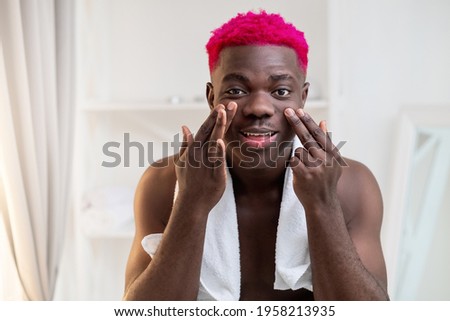 Facial care. Metrosexual black man. Beauty procedure. Face lifting. Morning grooming. Happy attractive shirtless african guy with bright pink hair towel on shoulder touching skin bathroom interior. Royalty-Free Stock Photo #1958213935