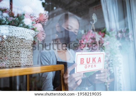 young woman florist wearing an apron and face mask standing by the window of a flower bucket giving an open text sign in the window